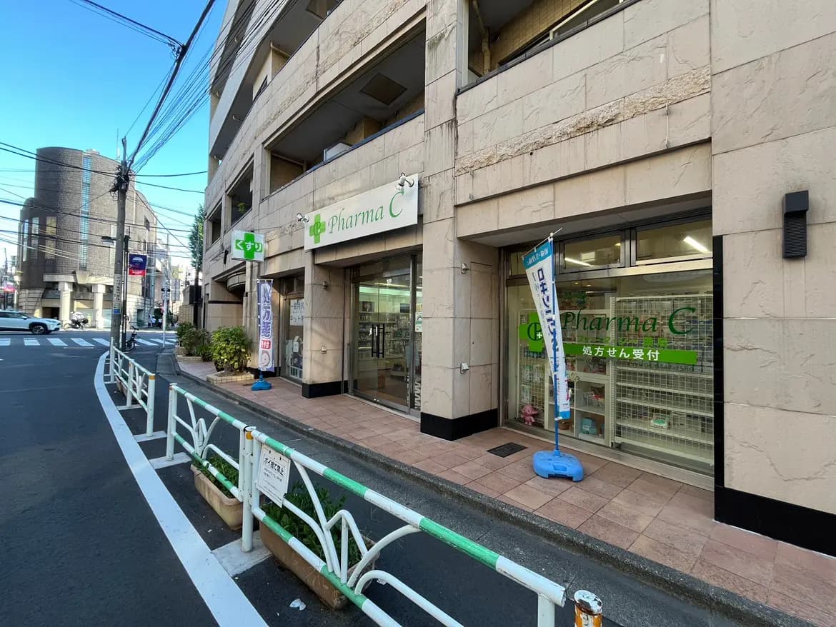 - 薬局「Pharma C」の外観、白と緑の看板、入り口には処方せん受付の案内、通り沿いの風景と青空の背景