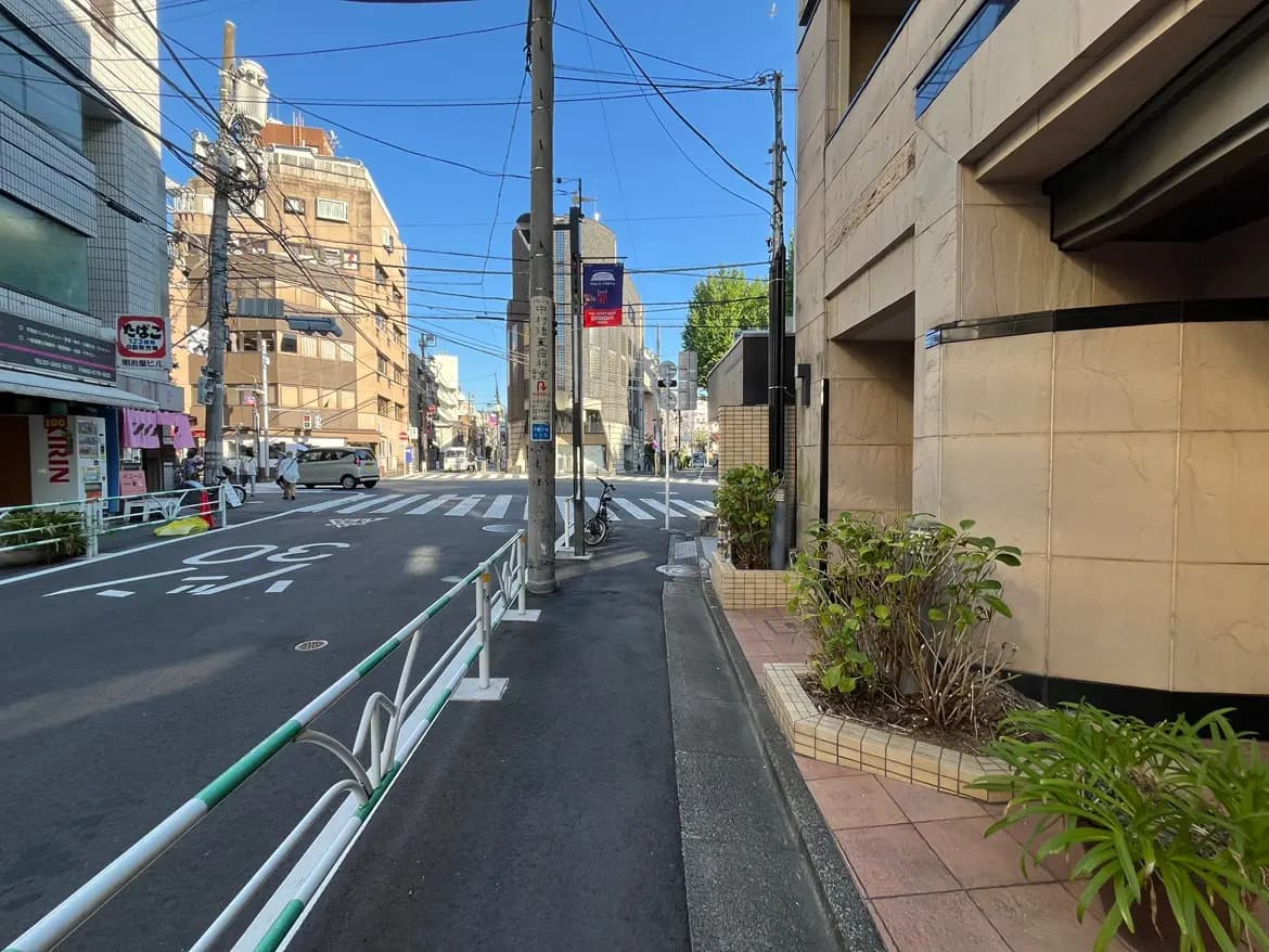 - 都会の街並み、青空の下での交差点と歩道、周囲に建物と電柱、植栽が見える風景