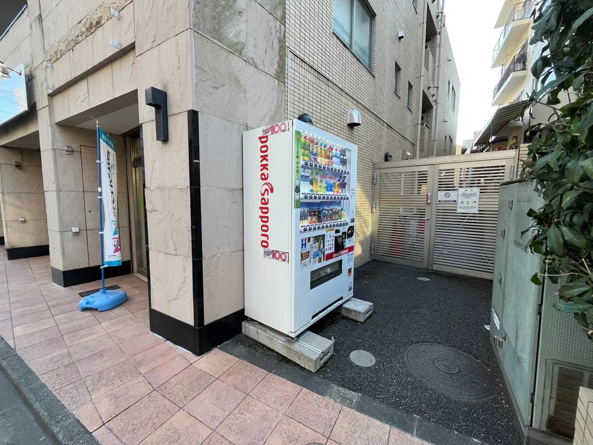 - 建物の入口横に設置された自動販売機、周囲にプランターと看板がある景観