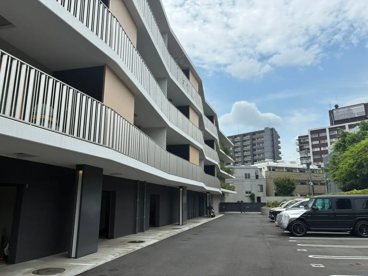 モダンなアパートメントビルの外観、曲線的なバルコニーと周囲に駐車場が広がる景観、青空と雲のある日。