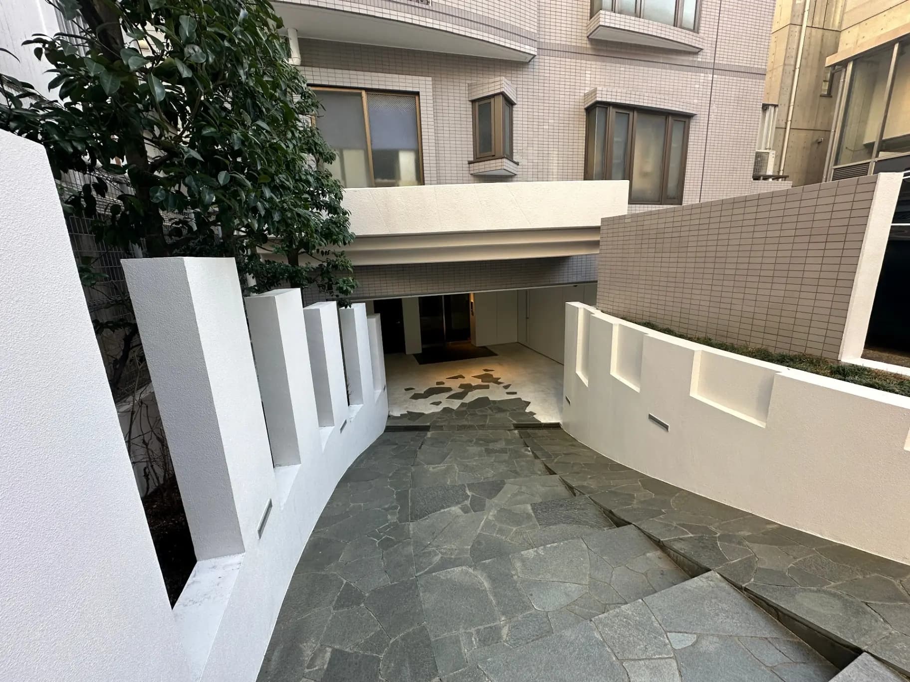 現代的な外観のマンションエントランス、白い壁と石畳のアプローチ、緑豊かな植物がある景観