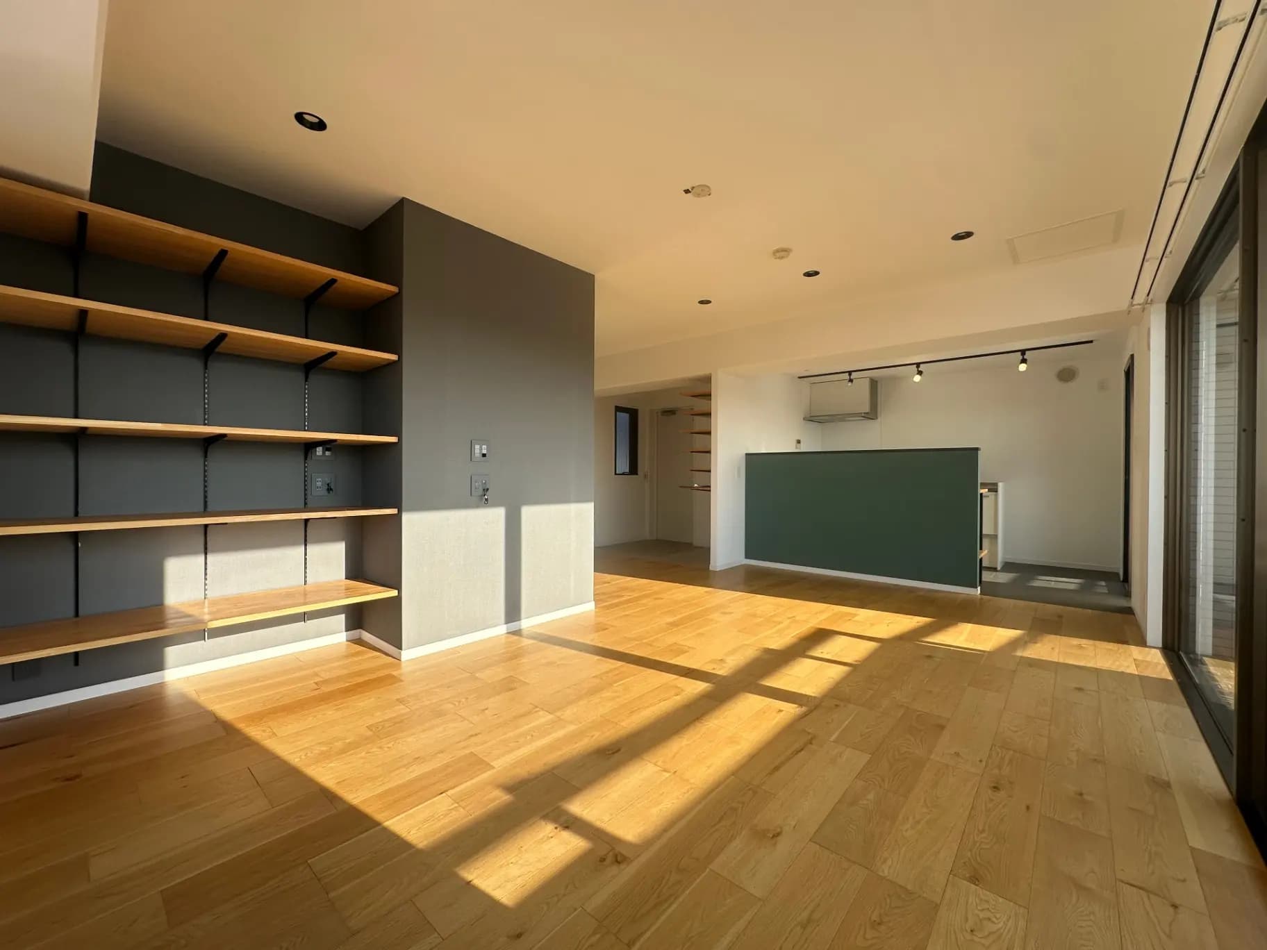 日差しが差し込むミニマリストのリビングルーム、木製のフローリングとグレーのアクセントウォール、収納棚とモダンな照明