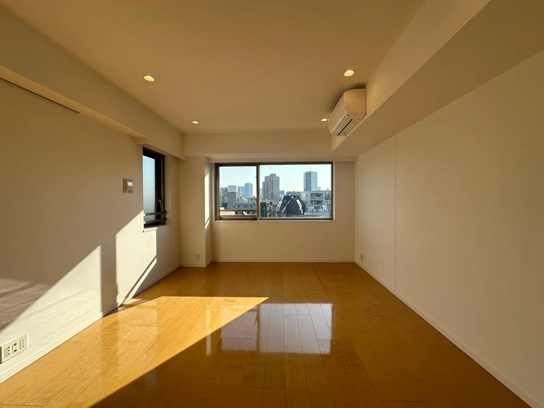 明るい自然光が差し込む広々としたリビングルーム、都市の景色が見える窓、木製フローリングのインテリア