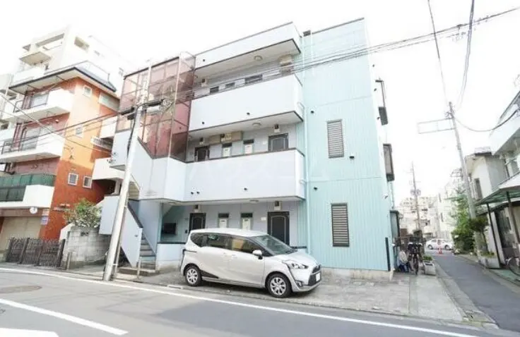 - 青い外壁の三階建てアパート、隣には赤い建物、道路脇に駐車された白いコンパクトカーのある風景