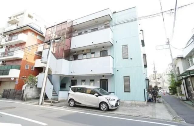 - 青い外壁の三階建てアパート、隣には赤い建物、道路脇に駐車された白いコンパクトカーのある風景