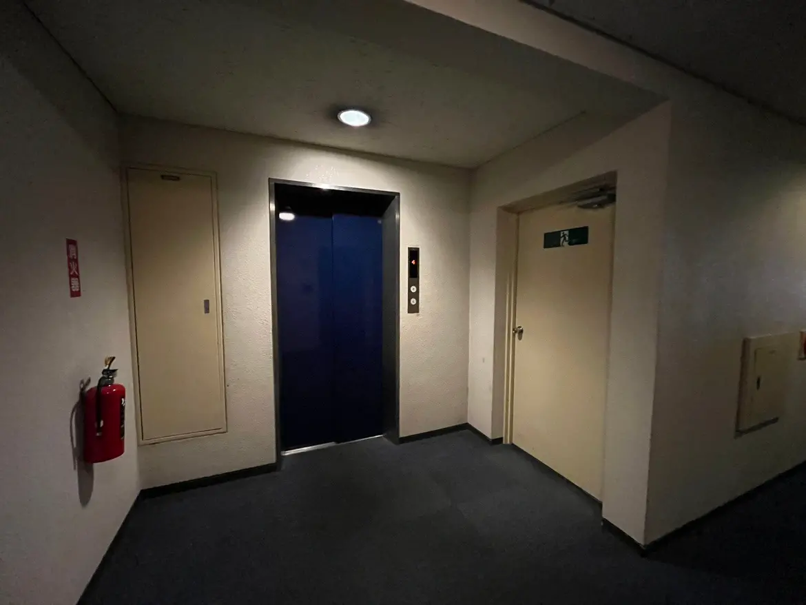 - エレベーターの前の廊下、左に消火器とその隣に収納スペース、右に非常口の扉がある室内の風景