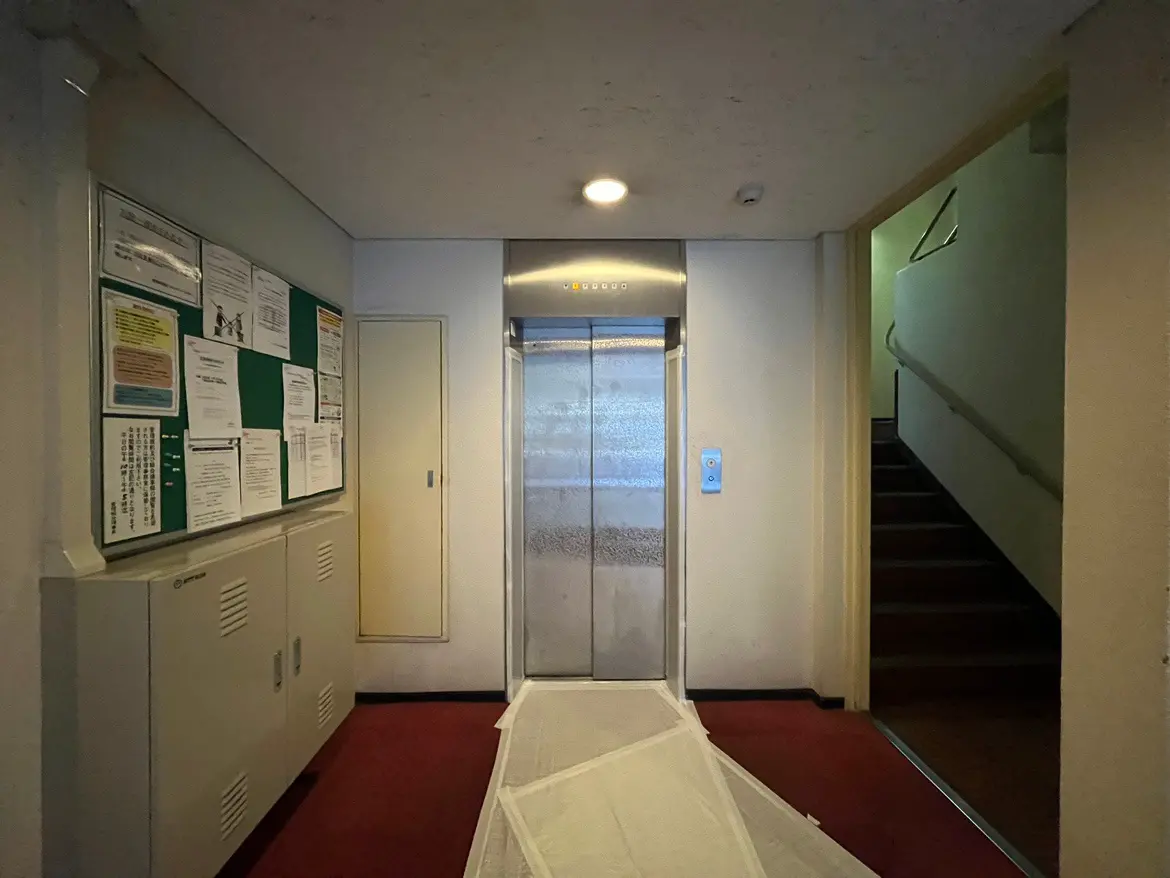 - エレベーターの前にある廊下、壁には掲示物があり、赤いカーペットと階段が見えるシンプルなインテリア