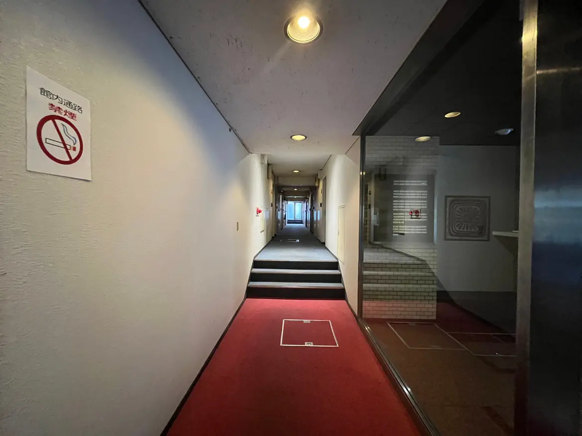- 赤いカーペットの廊下、禁煙の表示がある壁、行き止まりの明るい出口と階段のある長い廊下の風景