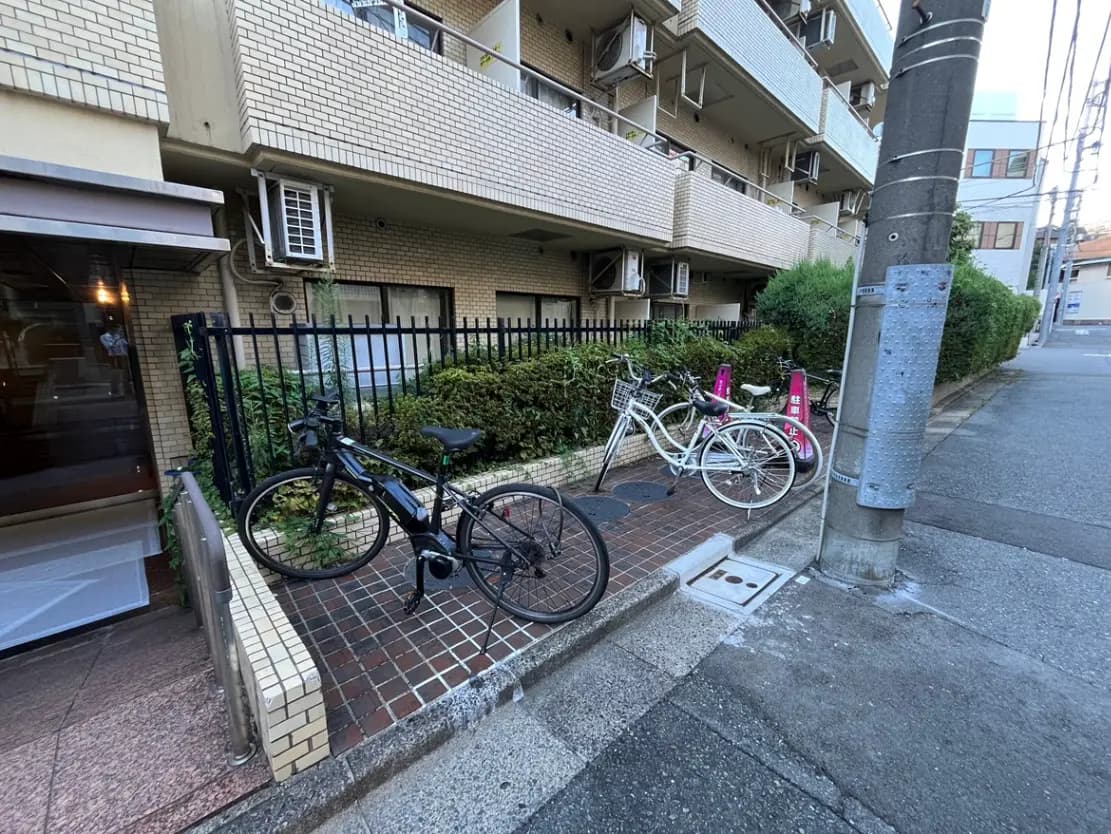 - アパートの前で停められた自転車、黒いスポーツバイクと白い自転車が並び、周囲には緑の植え込みとレンガの舗装がある風景