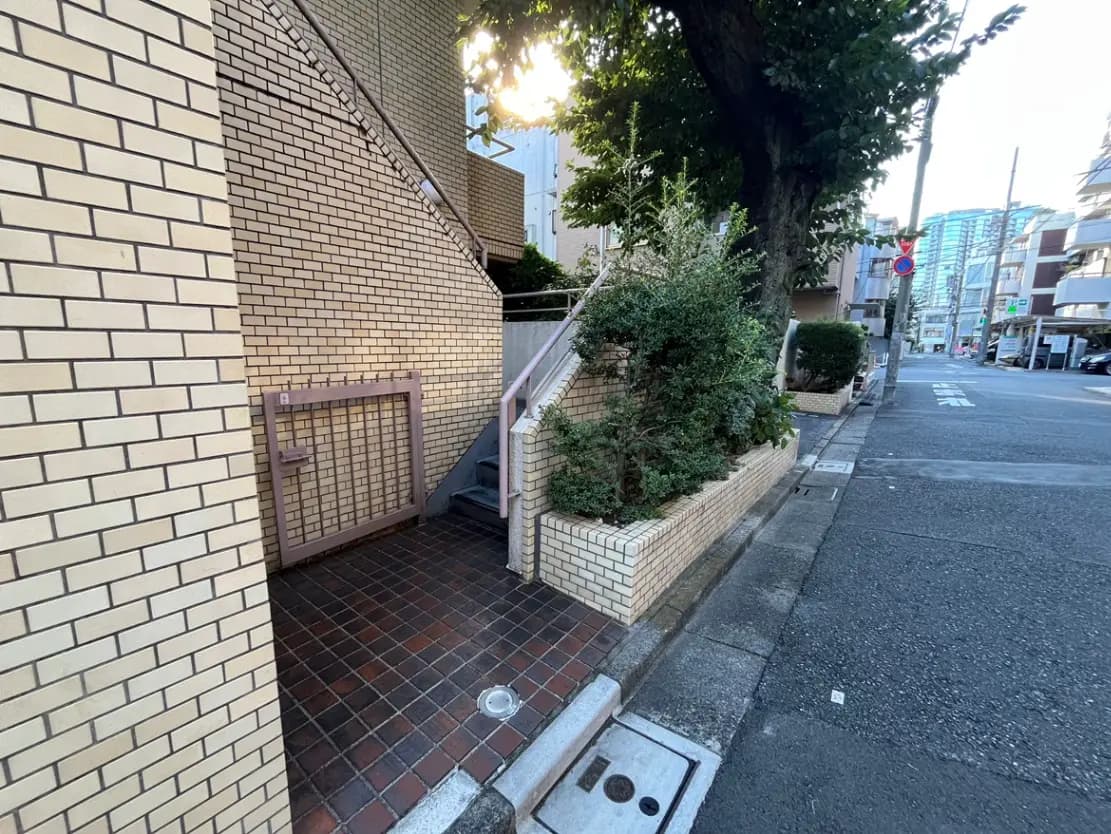 - 自宅のエントランスと階段、レンガの壁と植栽、明るい日差しが差し込む静かな通りの風景