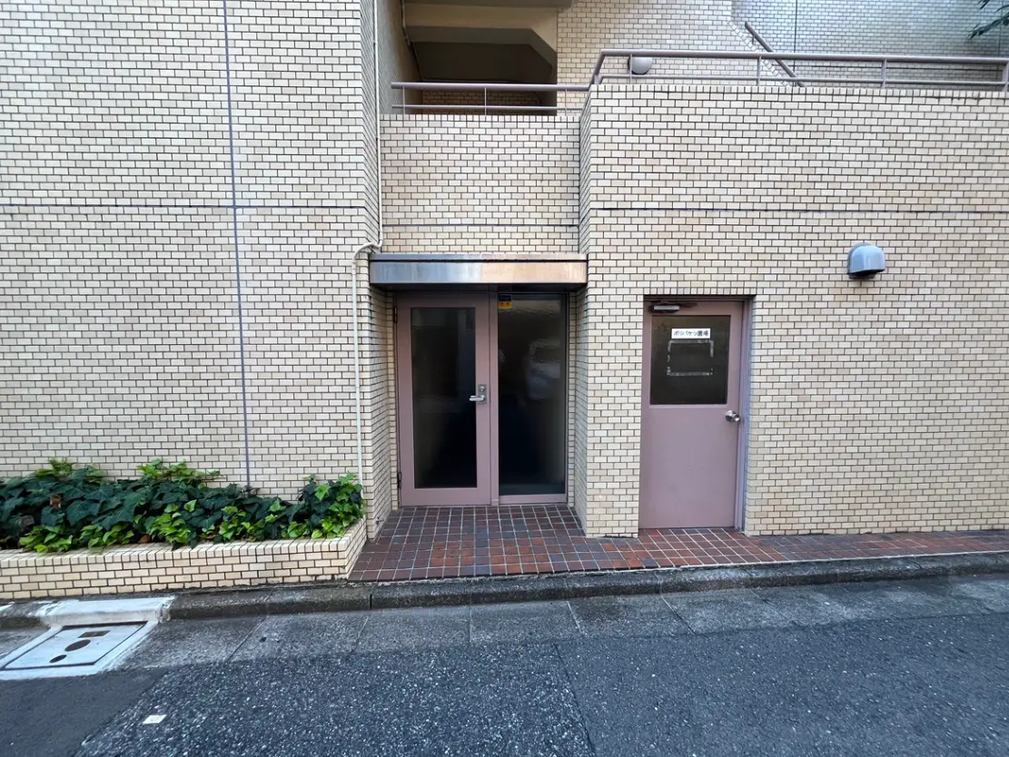 - 煉瓦タイルの外壁を持つ建物のエントランス、側に小さな窓付きのドアと緑の植栽がある景観