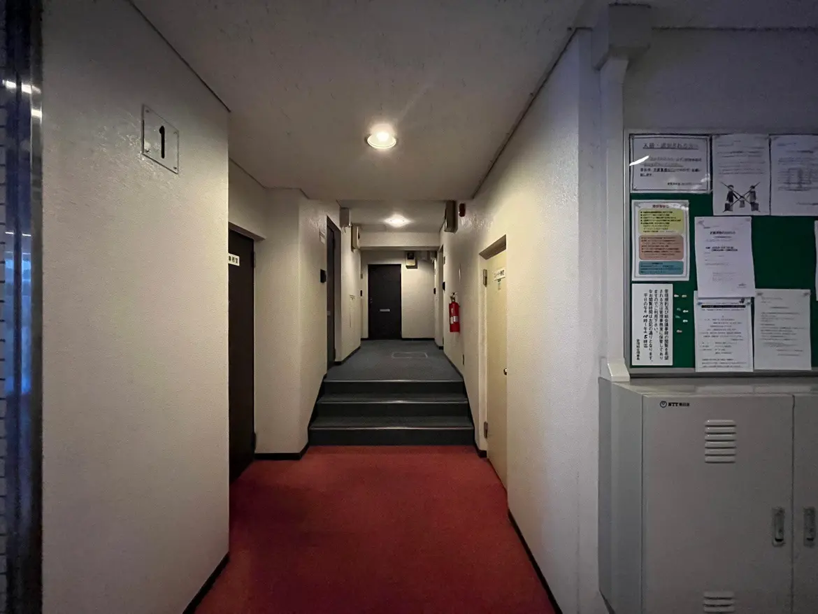 - 無地の壁と赤い絨毯が特徴的な廊下、左に番号の表示があり、右には掲示板とロッカーが配置されたインテリア