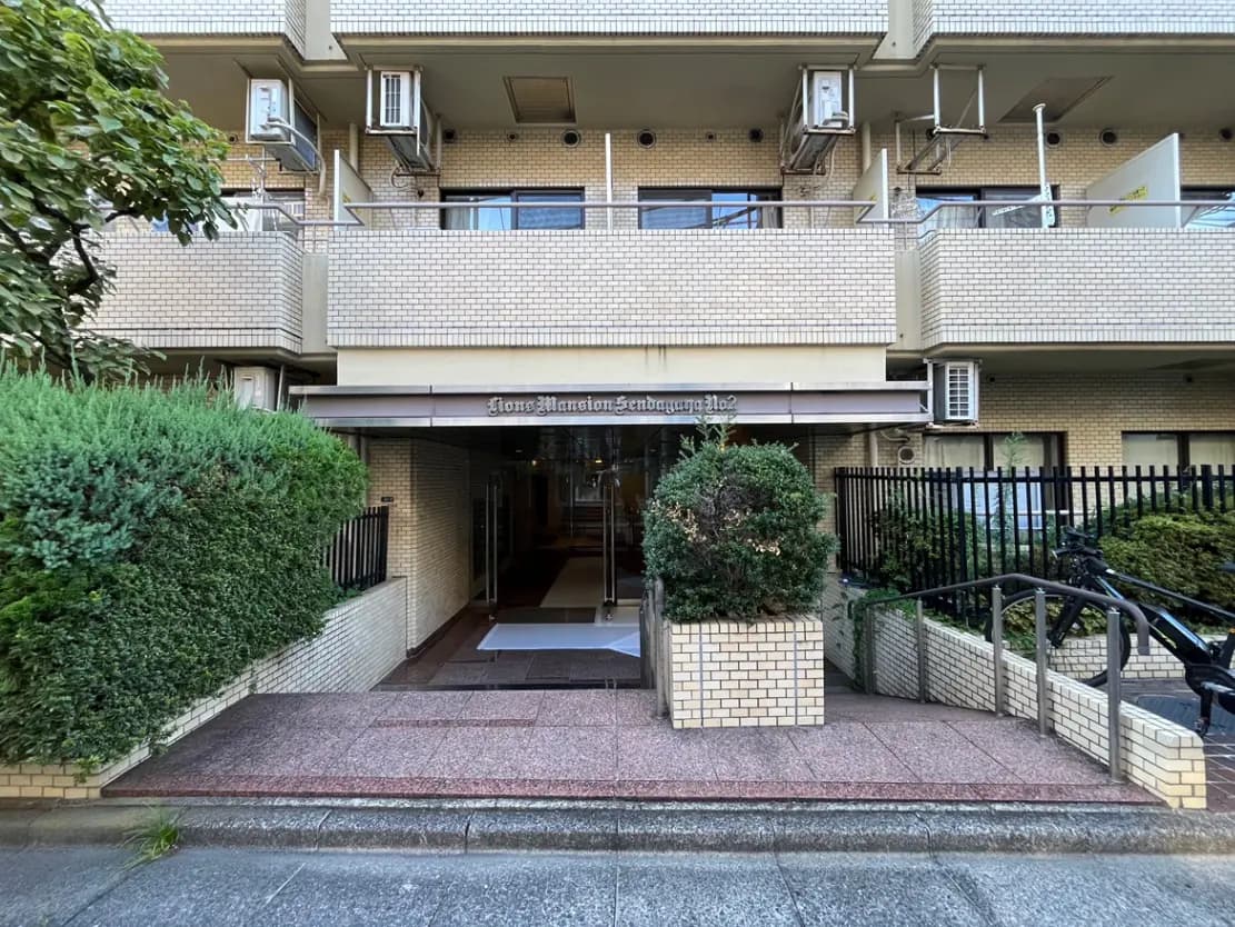 - スタイリッシュなマンションのエントランス、緑豊かな植栽と敷石のアプローチ、上部には窓が並ぶ外観