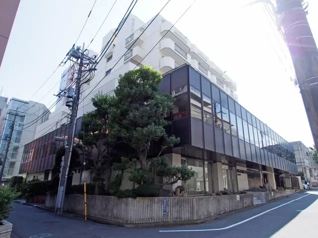 - 近代的なデザインの建物外観、ガラス張りのファサードと豊かな緑が調和した景観、周辺には街路樹と電柱が見える街並み