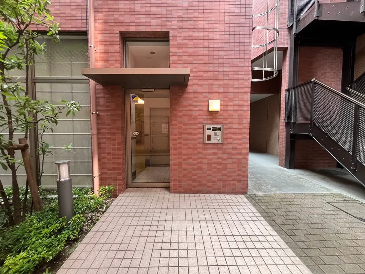 - ブロックタイルの外壁と植物に囲まれた建物の入り口、明るい照明と呼び鈴が設置されたエントランスの様子