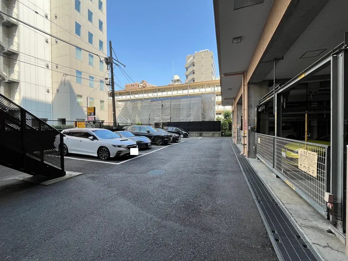 - 駐車スペースに停まっている複数の車と周囲の建物、明るい青空の下での様子。
