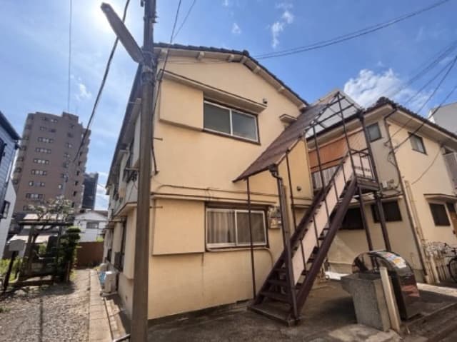 - 住宅の裏側、階段付きの外観と周囲の建物、晴れた日の明るい空が映えるシーン