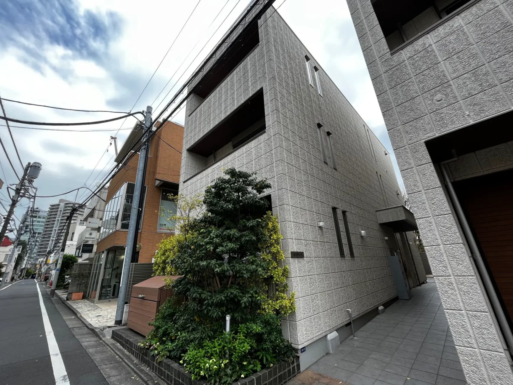 都会の通り沿いにあるモダンな外観の建物、グレーの壁と窓、緑の植栽が引き立つ景観