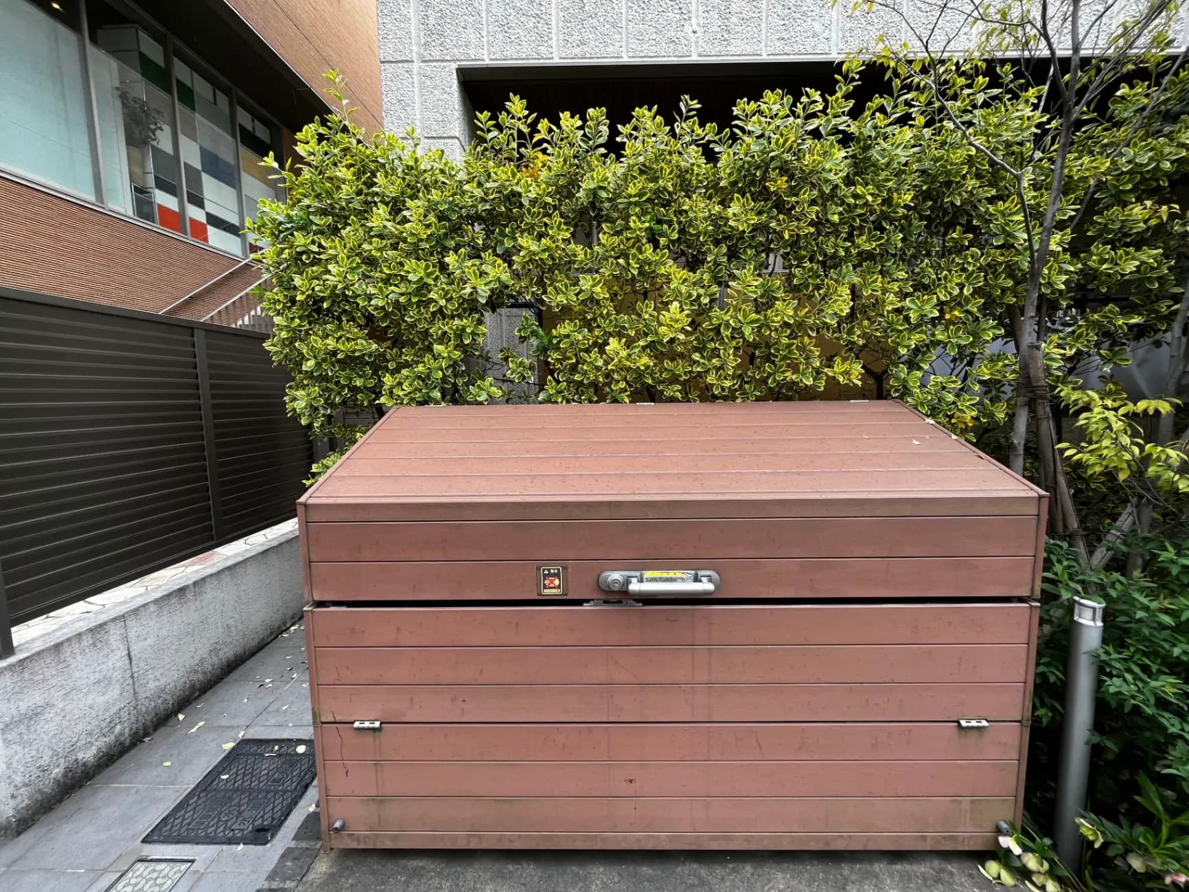 茶色の屋外収納ボックス、背景に緑の植栽とモダンな建物の外観