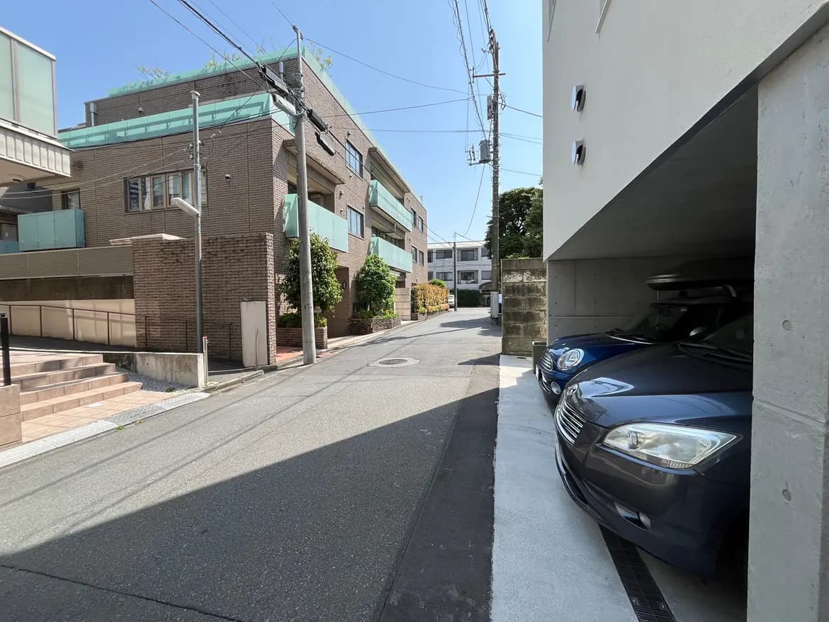 - 住宅街の静かな通り、コンクリート製の駐車場に停まる青い車と周囲のマンション、青空が広がる風景