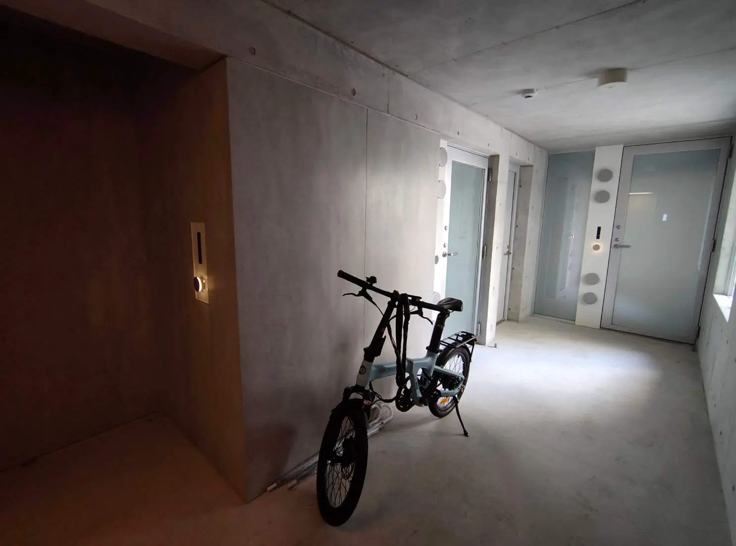 - モダンなコンクリート造の廊下に置かれた折りたたみ自転車、ガラスドアと照明スイッチのあるシンプルなインテリア