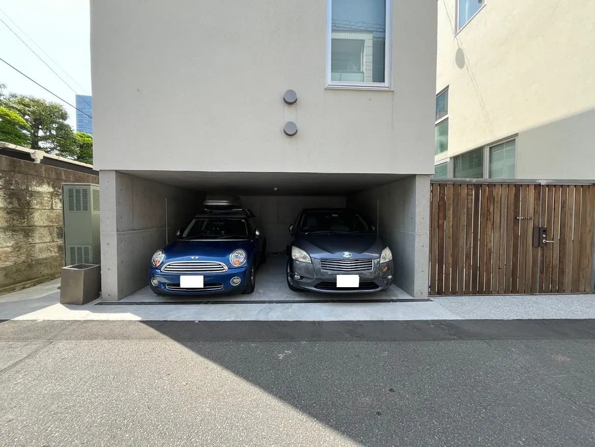 - モダンな住宅の車庫、青いミニクーパーとグレーのセダンが駐車されている様子、周囲にはコンクリートの壁と木製のゲートが見える