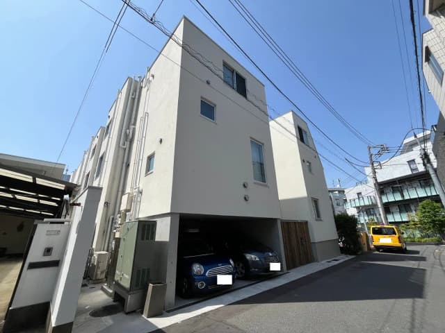 - 青空の下に立つ現代的な住宅、ガレージに停車中の車、周囲の電線とアスファルトの道路が見える景観