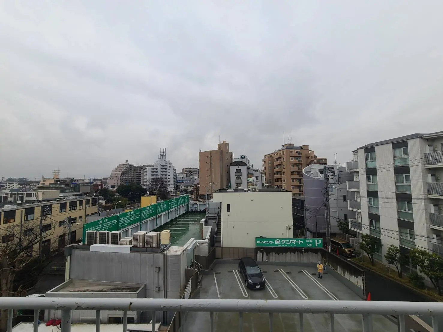 都市の風景、雲が多い曇天の下、ビルやマンションが並ぶ街並み、駐車場と歩行者が見える