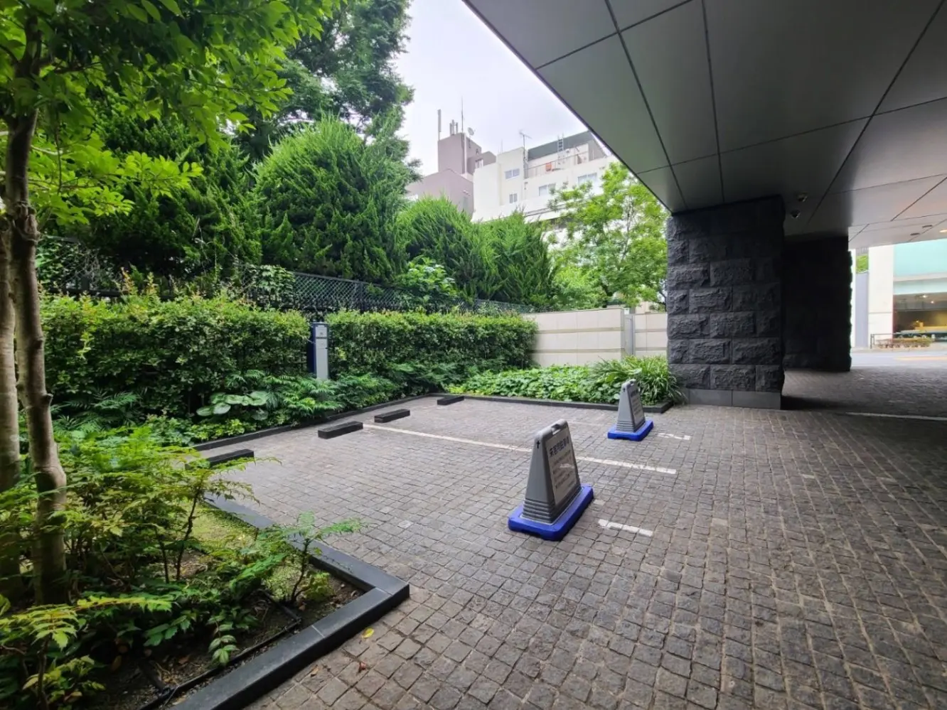 都市のビルに囲まれた緑豊かな庭園と駐車スペース、石畳の歩道と植栽が美しい外観