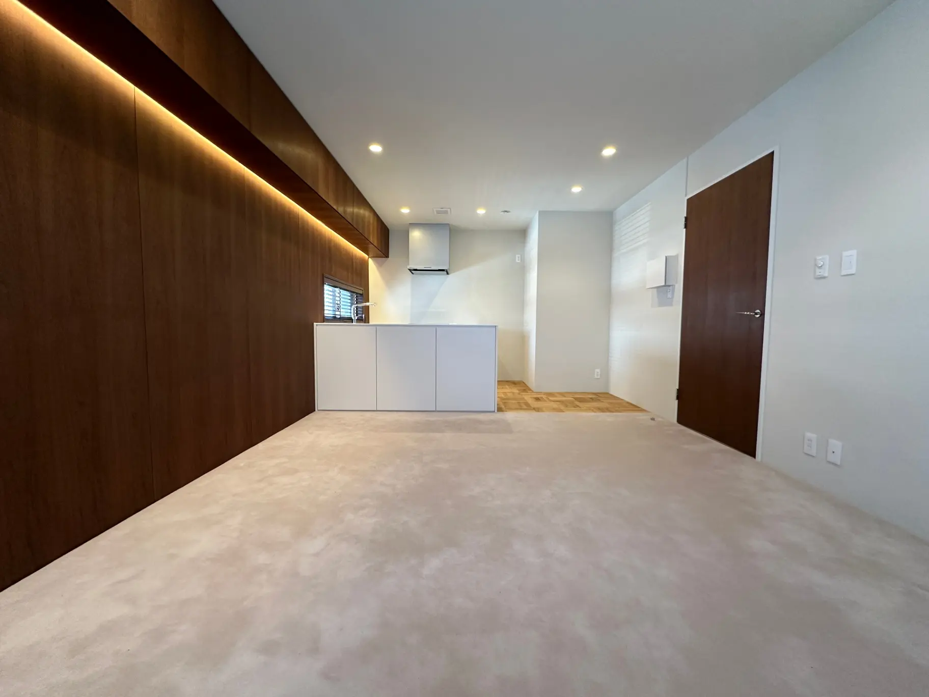 モダンなデザインの広々としたリビングルーム、木目の壁パネル、シンプルなキッチンキャビネット、埋め込み型照明で明るい空間