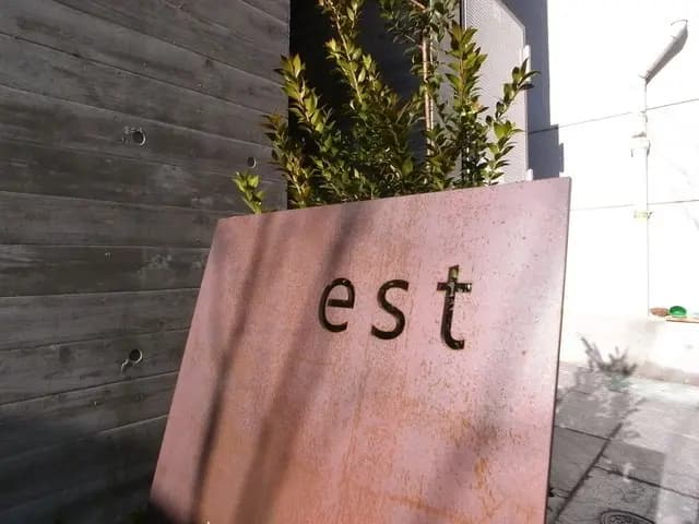 錆びた銅製の看板に刻まれた「est」の文字と緑の植物が映える外観デザイン