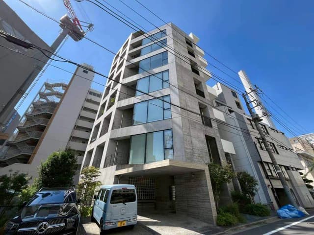 - モダンなコンクリート製のマンション、青空を背景にした外観、駐車スペースに停まる車と周囲の建物が映る写真