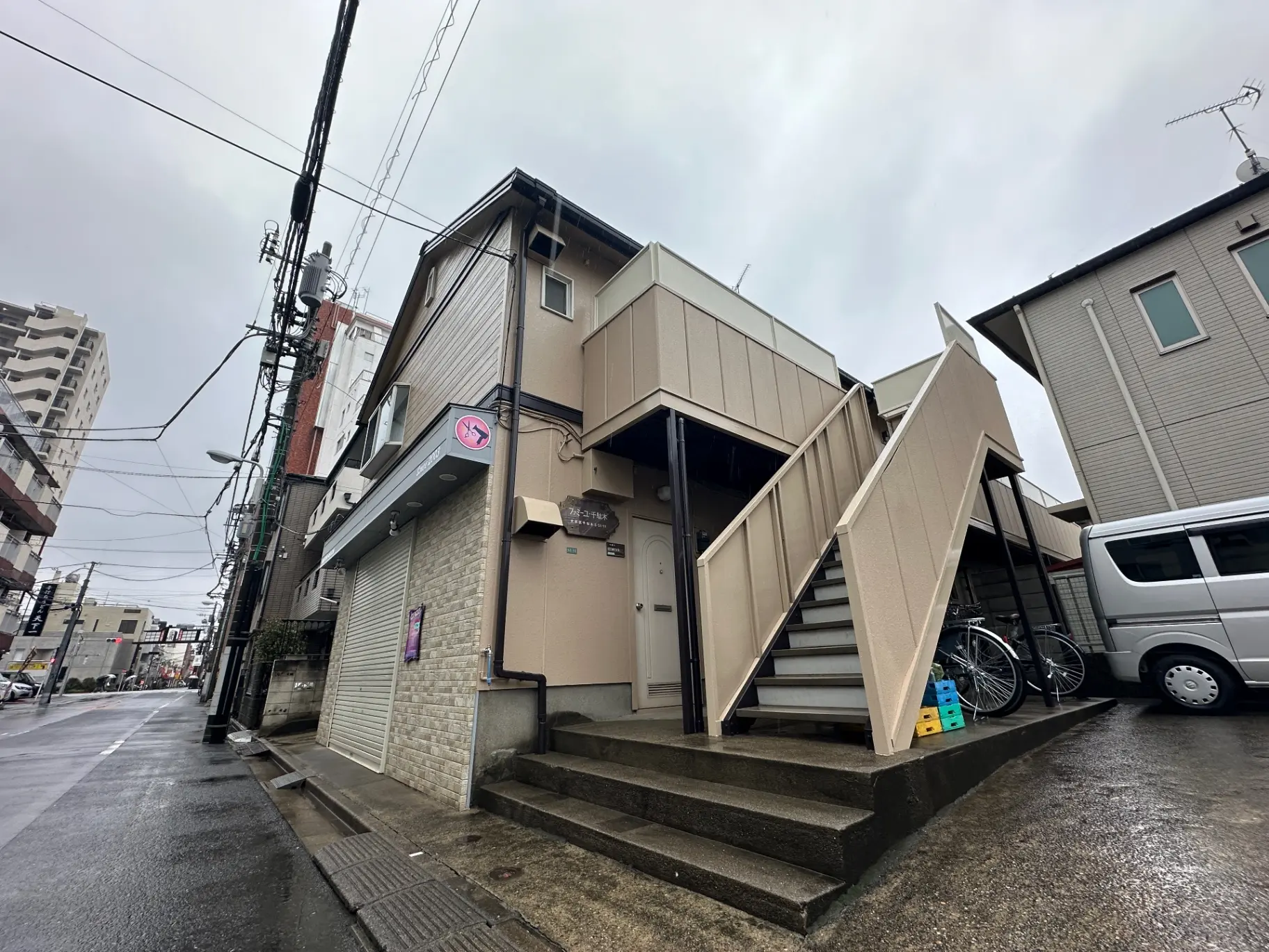 日本の都市部の狭い通りにあるアパートメントの外観、雨の日の曇り空と路上駐車の車両