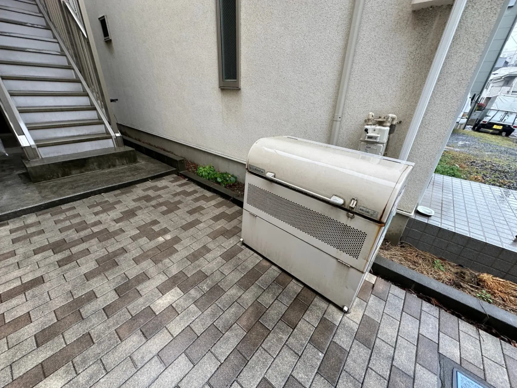 アパートのエントランス付近の模様付きタイル敷きの広場とゴミ箱、階段と外部配管が見える外観