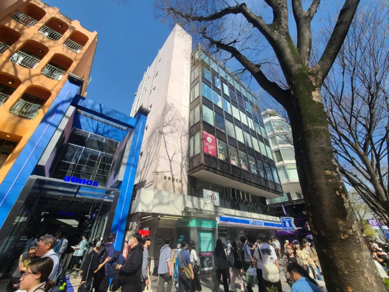 明るい日差しの下、多くの人々が行き交う都市の通り、特徴的な建物とファッションショップのロゴが目立つ風景