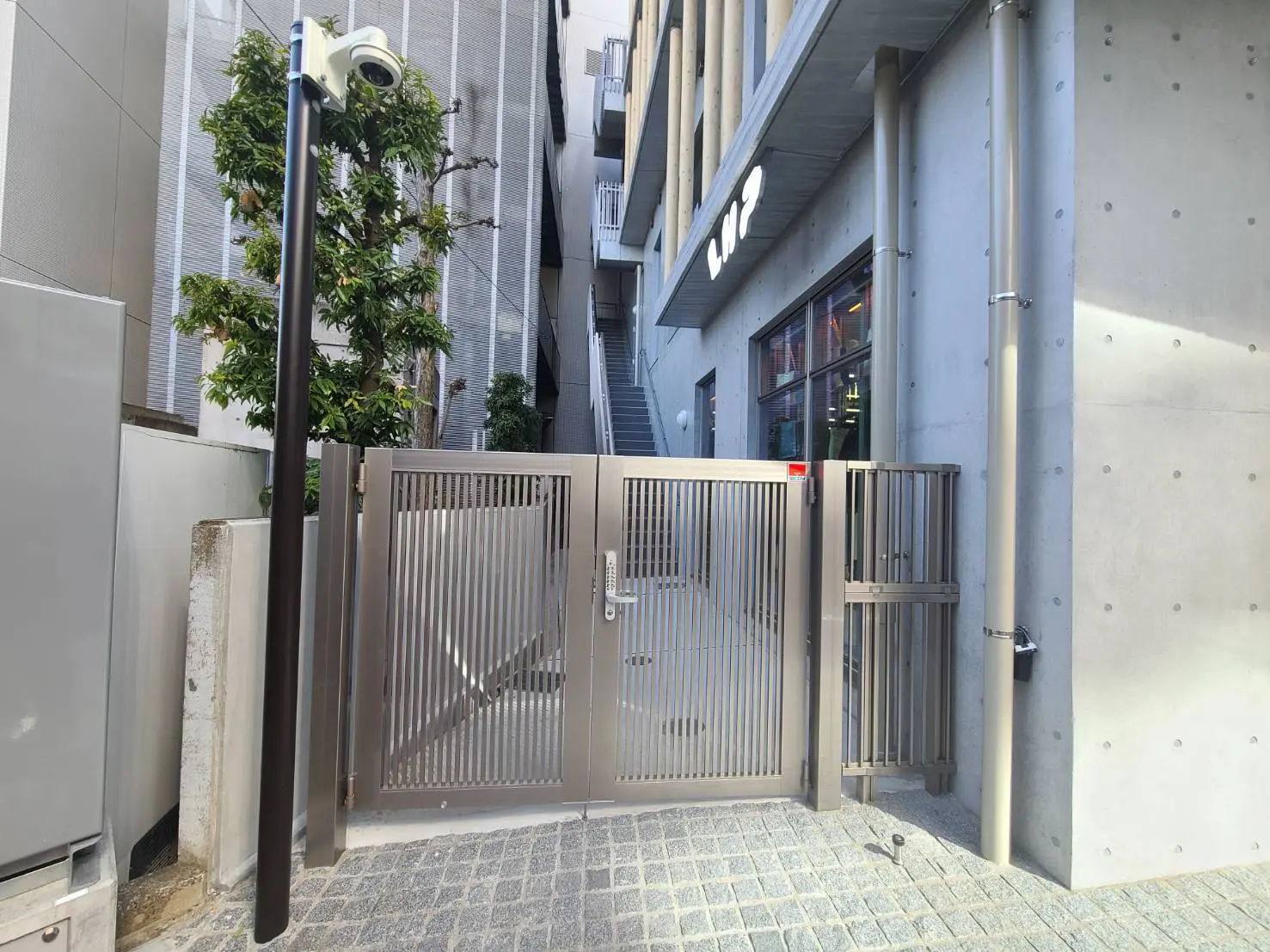 都市の建物入り口、モダンなデザインの鉄製ゲートとコンクリート壁、防犯カメラ付き