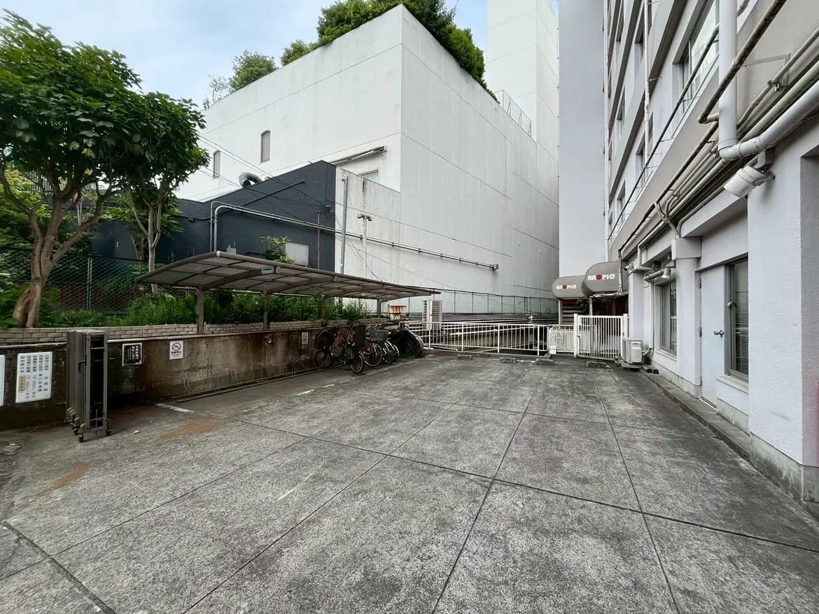 - 住宅の裏庭スペース、自転車スタンドと緑の木々、近隣の建物の壁が見える景観