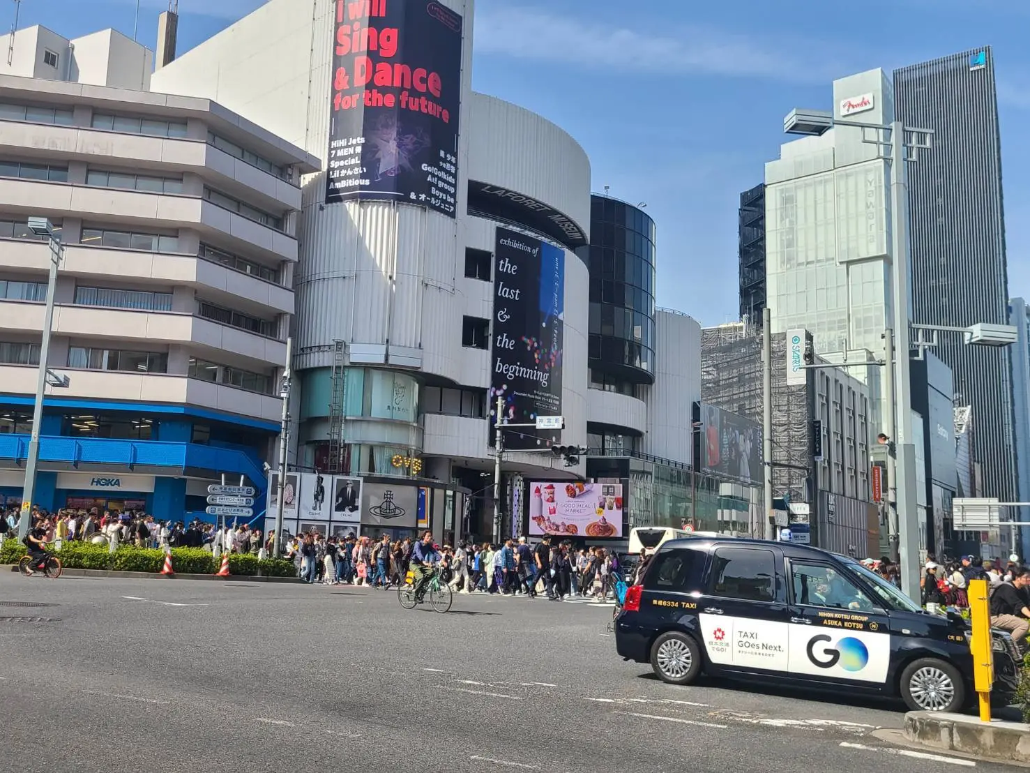 東京の繁華街、人混みが多い交差点、ビルの大型広告看板、タクシーと自転車が通る都市風景