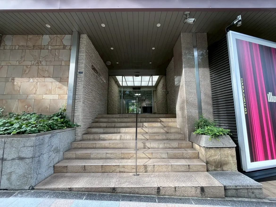 - エントランスの階段、モダンな外壁と緑の植栽がある建物入り口、透明な屋根からの自然光が差し込む景観