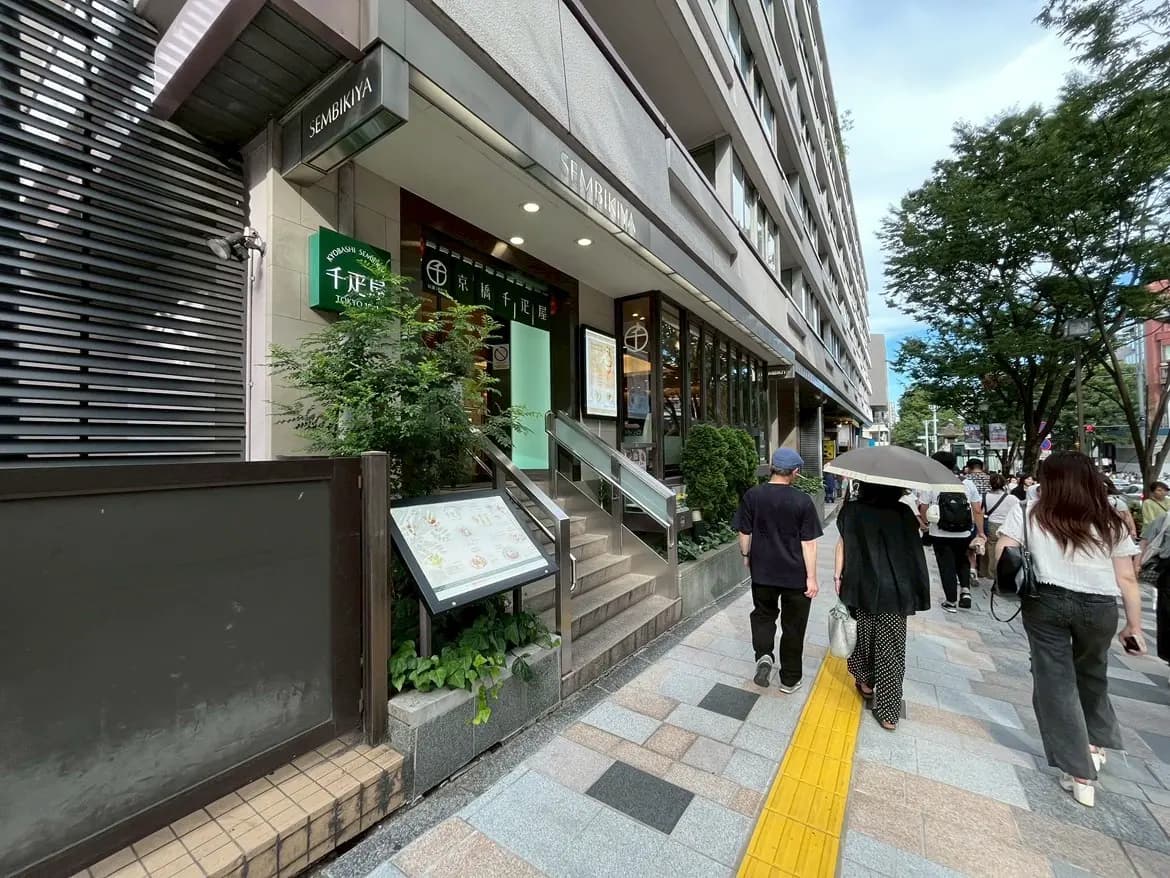 - 商業ビルの入口と歩道、緑豊かな植栽と人々が行き交う様子、晴れた日の下での街の風景