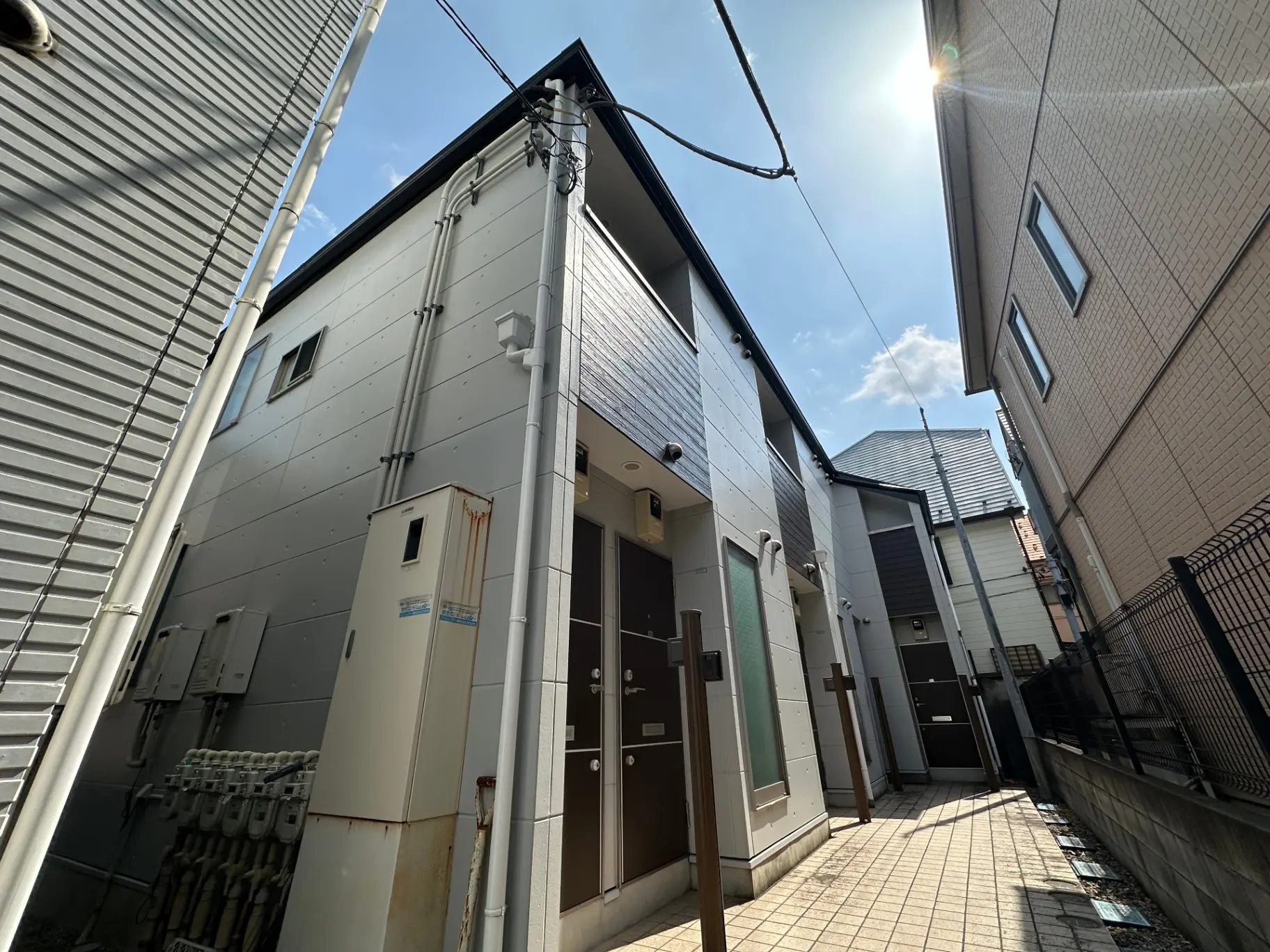 日差しの強い日本の住宅街、狭い路地に建つ現代的なデザインの家、グレーとベージュの外壁