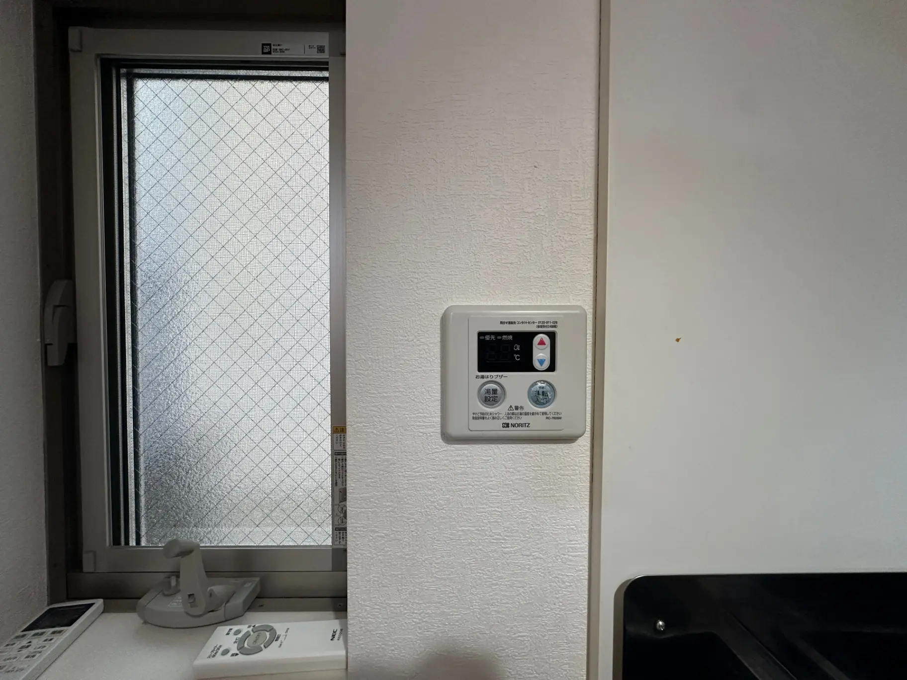 キッチンの壁に取り付けられたノーリツの給湯器リモコン、防犯ガラスの窓と白い壁のインテリア