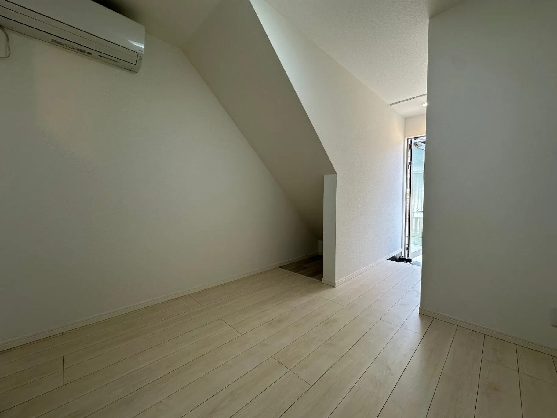 シンプルなデザインの居間、明るい照明と薄色木製のフローリング、傾斜のある天井と扉付きのバルコニー