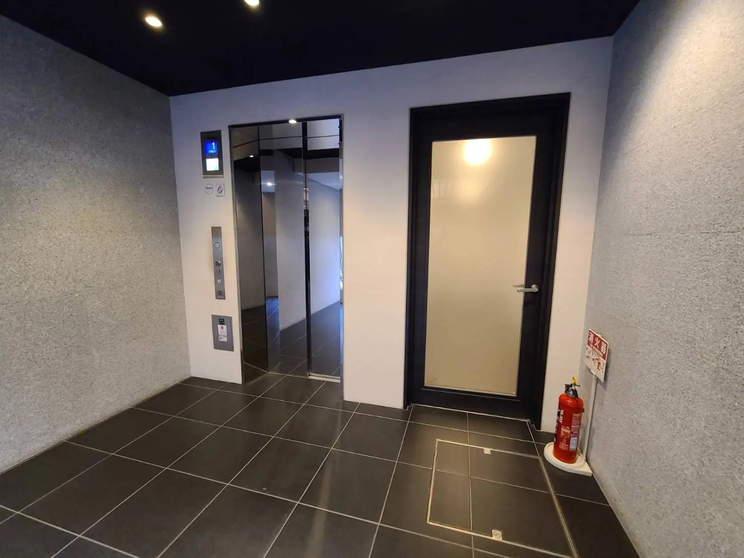 モダンなデザインのエレベーターホール、黒いタイルの床とグレーの壁、消火器とインジケーター付きのエレベーターと白いドア