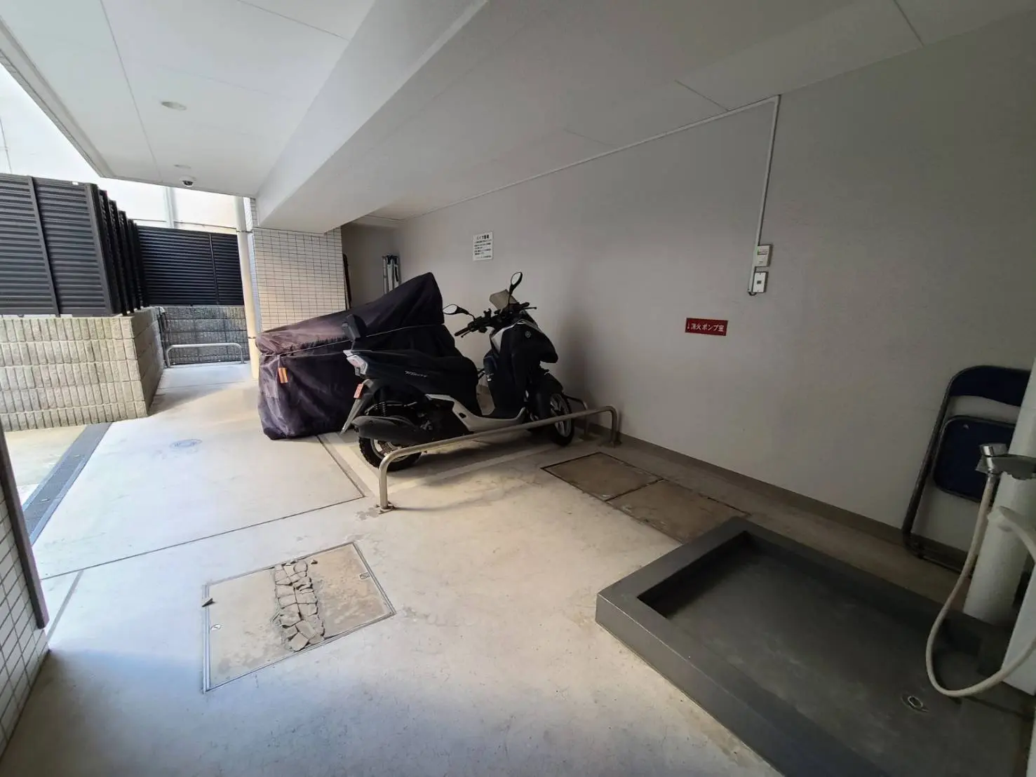 屋外のバイク置き場、バイクカバーがかかったスクーターと灰色の床、建物の壁に掲示された「放火対策」の看板