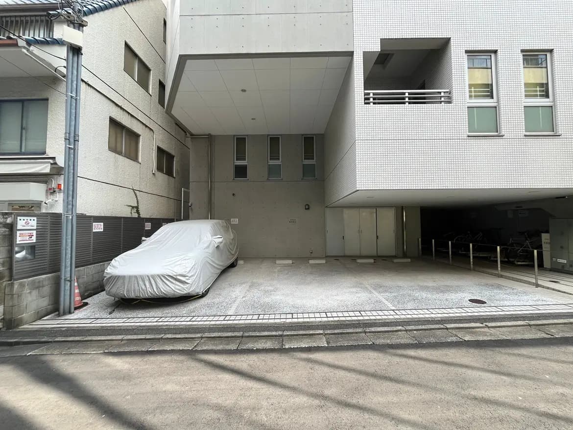 - 駐車場にカバーをかけた車が停まっている、周囲にコンクリート製の壁と住宅があるスペース