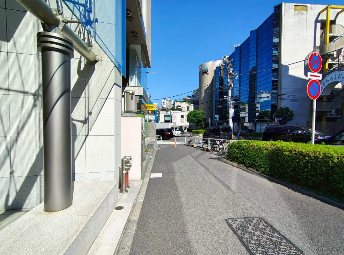 - 青空の下、都会的な景観を背景にした舗装された歩道と建物側面、手前に配置されたガーデンと道路の様子