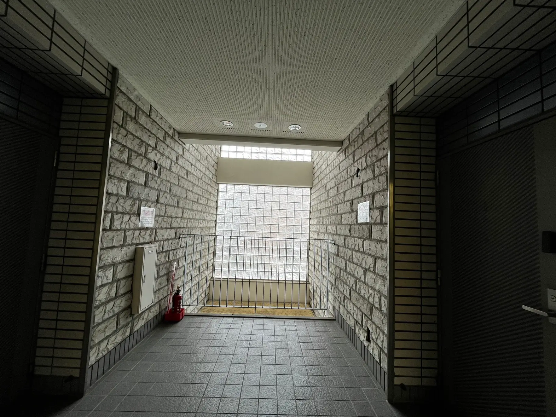 モダンなマンションの共用エントランスホール、自然光が差し込む大きな窓、灰色のタイル床と白いレンガ壁のデザイン
