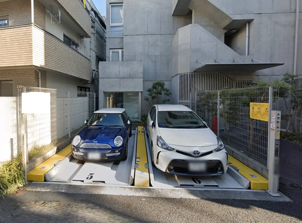 - 住宅の駐車場に停められた青いミニクーパーと白いトヨタプリウス、周囲にはコンクリートの壁と緑の植栽があるエリア
