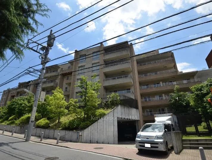 - 青空を背景にしたモダンなマンションの外観、周囲には緑豊かな植栽と駐車スペースがある風景
